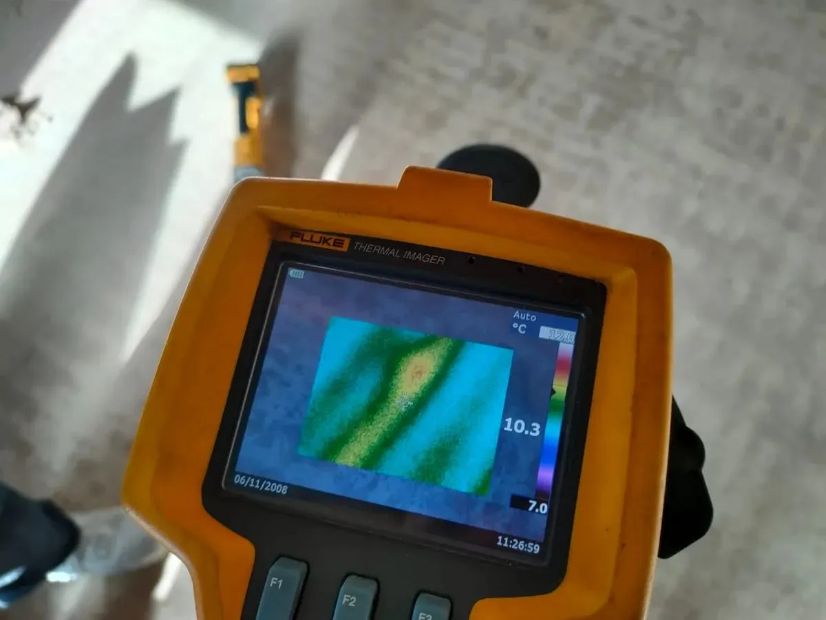 Handheld thermal camera screen showing warm pipe lines across a cold screed floor